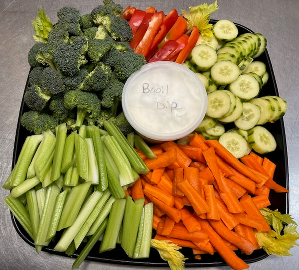 A platter of fresh vegetables including broccoli, carrots, cucumbers, and zucchini, with a container of dip.